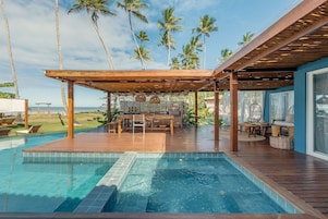 Una terraza de madera con piscina, mobiliario exterior y vistas al océano y palmeras.