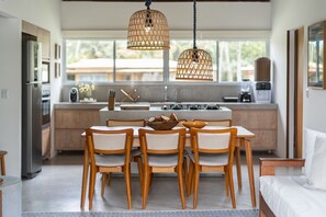 A modern kitchen with a dining area, featuring a central island, wooden chairs, and hanging wicker light fixtures. - Orixás Residence (Cairu)