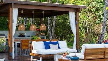 Un patio al aire libre con piso de madera, asientos blancos acolchados, lámparas de mimbre colgantes y una cocina al fondo.