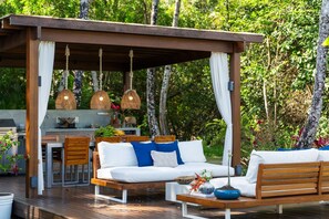 An outdoor patio with a wooden deck, white cushioned seating, hanging wicker lamps, and a kitchen area in the background. - Orixás Residence (Cairu)
