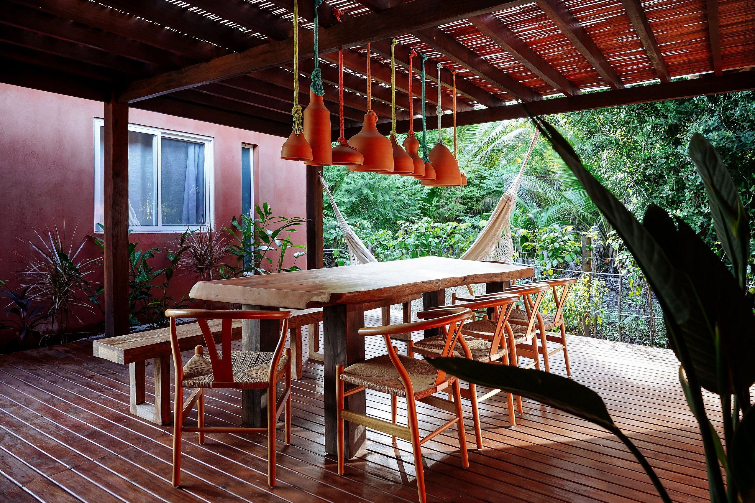 Un terrazzo in legno con tavolo e sedie, circondato da piante tropicali e un edificio rosso sullo sfondo.