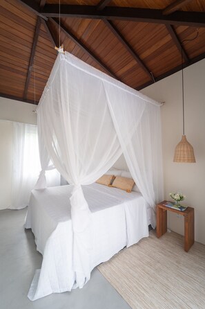 A bedroom with a canopy bed, wooden ceiling beams, a woven side table, and a hanging light fixture. - Orixás Residence (Cairu)