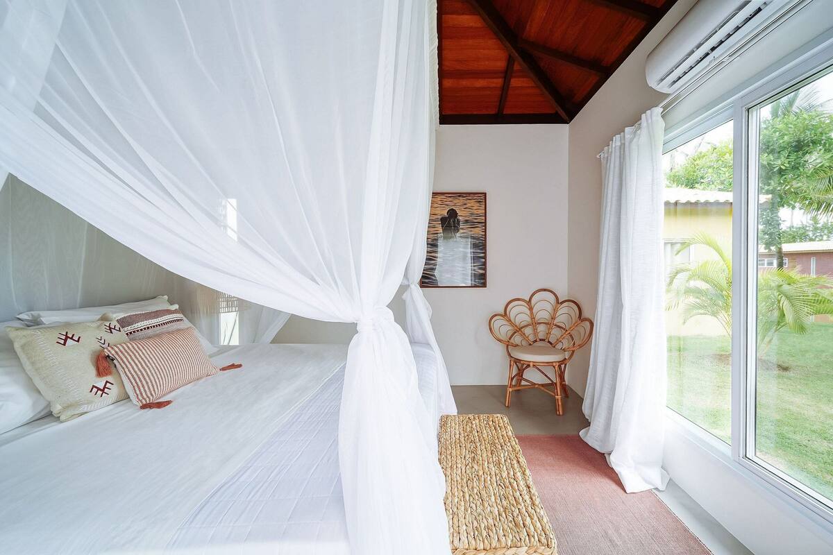 A bedroom with a bed, white curtains, a wicker bench, and a view of greenery outside.