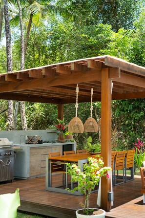 Uma área de cozinha ao ar livre com churrasqueira, mesa de jantar e cadeiras, cercada por vegetação exuberante e uma pérgula de madeira.