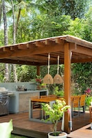Uma área de cozinha ao ar livre com churrasqueira, mesa de jantar e cadeiras, cercada por vegetação exuberante e uma pérgula de madeira.