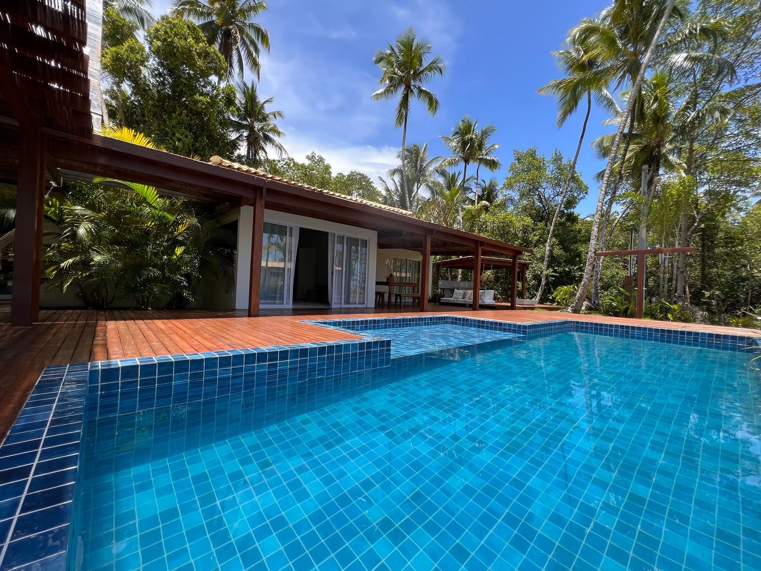 A swimming pool with tiled edges, surrounded by a wooden deck and a covered area with seating.