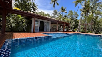 A swimming pool with tiled edges, surrounded by a wooden deck and a covered area with seating.