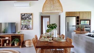 A dining area with a wooden table set for four, surrounded by wicker chairs, a hanging light fixture, and a television on a wooden stand.