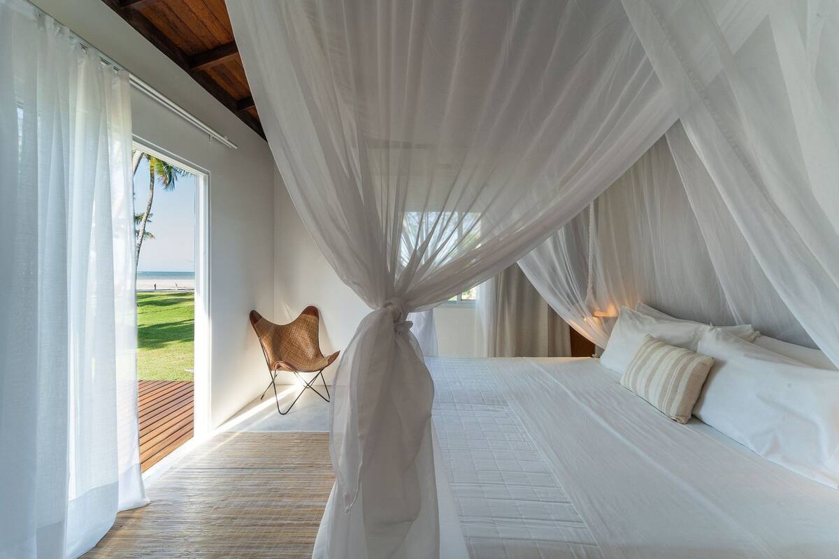 A bedroom with a bed, white curtains, a chair, and a view of the beach.