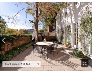 Un giardino anteriore con un tavolo rotondo e sedie, circondato da un muro in mattoni e un edificio bianco.