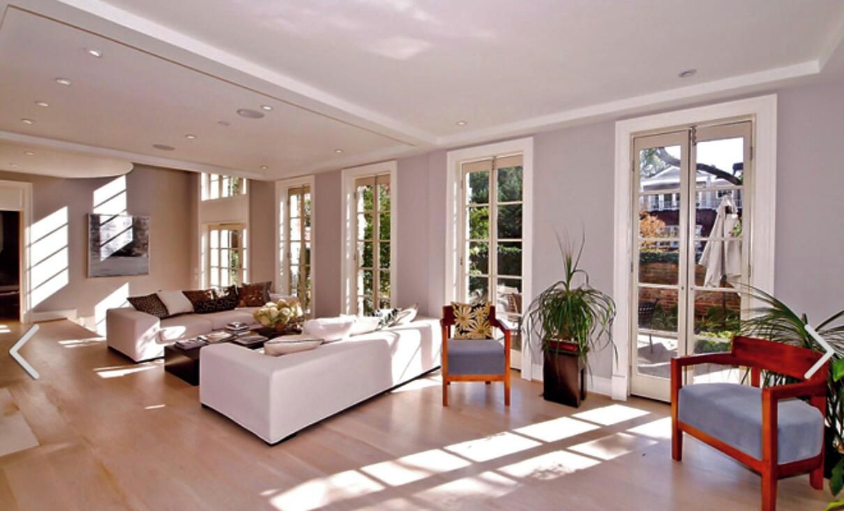 A spacious living room with a large sofa, two armchairs, and a coffee table. There are large windows allowing natural light and a view of the outdoors.