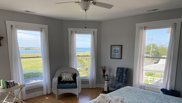 Quarto com cama, duas cadeiras, uma mesinha e amplas janelas com vista para o exterior.