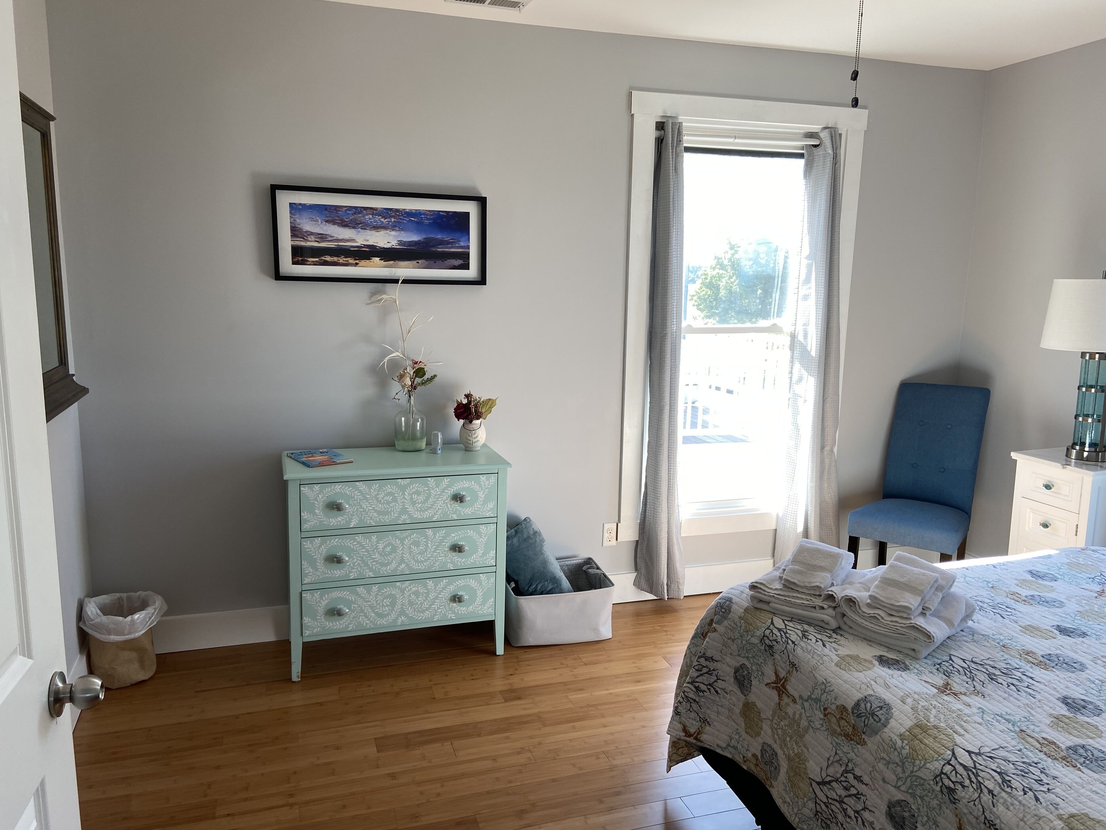 A bedroom with a bed, a dresser, a chair, a window with curtains, and a framed picture on the wall.