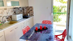 A compact kitchen with a microwave, sink, and a small stove. There is a table with a few red chairs and a glass top. A balcony with a green railing is visible through an open door.