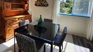 A dining area with a glass-top table, patterned chairs, a wooden cabinet, and a view of trees through a window. - Asian-themed Queen's Loft Apartment 10 mins to the beach and downtown! (GUALALA)