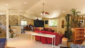 A spacious living room with a red sofa, a white coffee table, and a ceiling fan. There is a large window providing natural light and a view of the outdoors. - Asian-themed Queen's Loft Apartment 10 mins to the beach and downtown! (GUALALA)