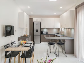 A modern kitchen with a dining area, featuring a built-in oven, refrigerator, and a flat-screen TV mounted on the wall. - Fully renovated apartment in the city centre of Chania with a roof garden! (Χανιά)