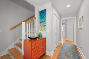 A staircase with wooden handrails, a red dresser with drawers, a colorful abstract painting, and a patterned rug on a hallway with white walls and wooden flooring. - Chatham Luxury Retreat—Porch, Art & Family Fun (Chatham)
