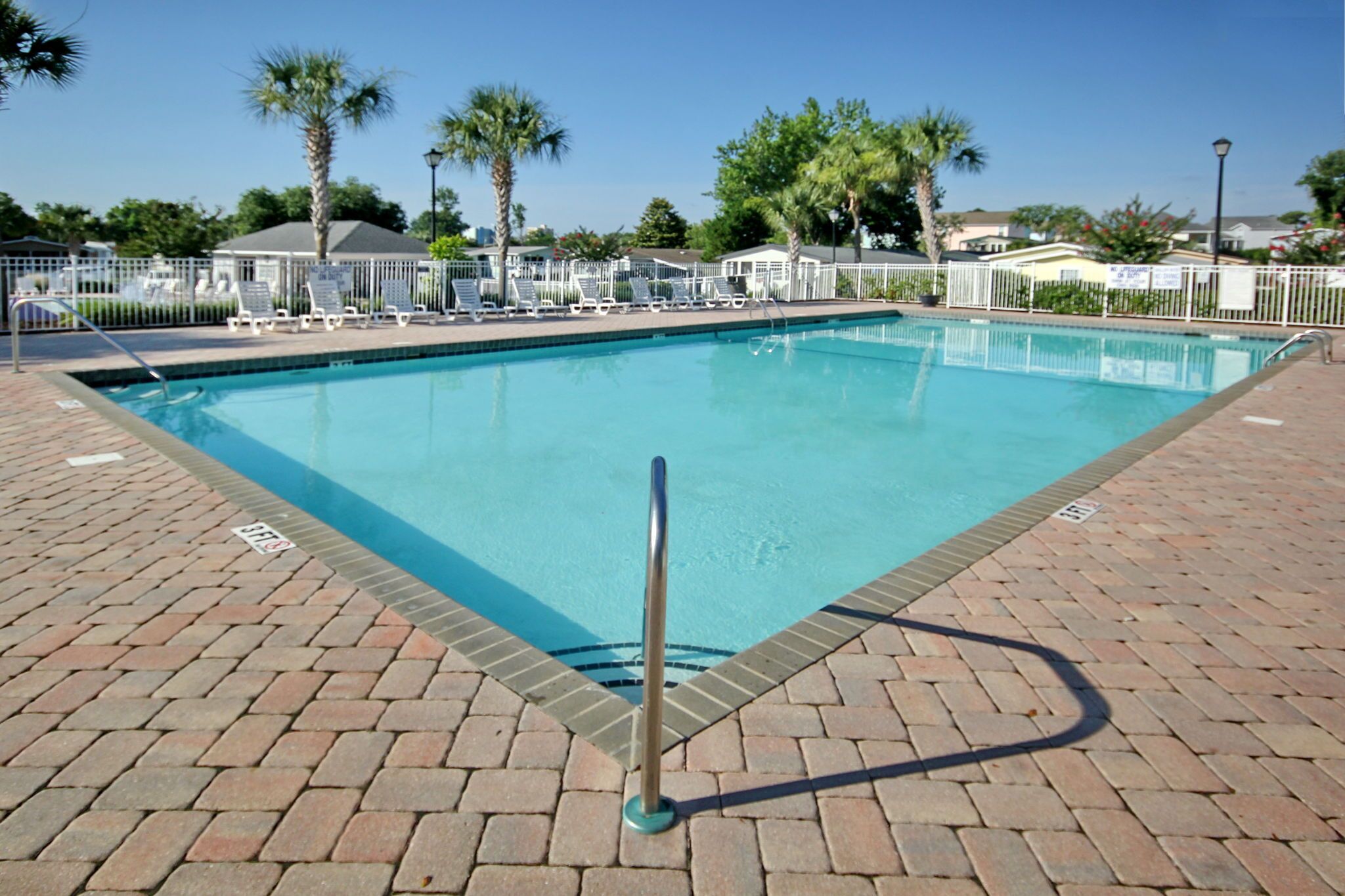 Une piscine rectangulaire dotée d’une barrière de sécurité en métal, entourée d’une clôture blanche et de palmiers.
