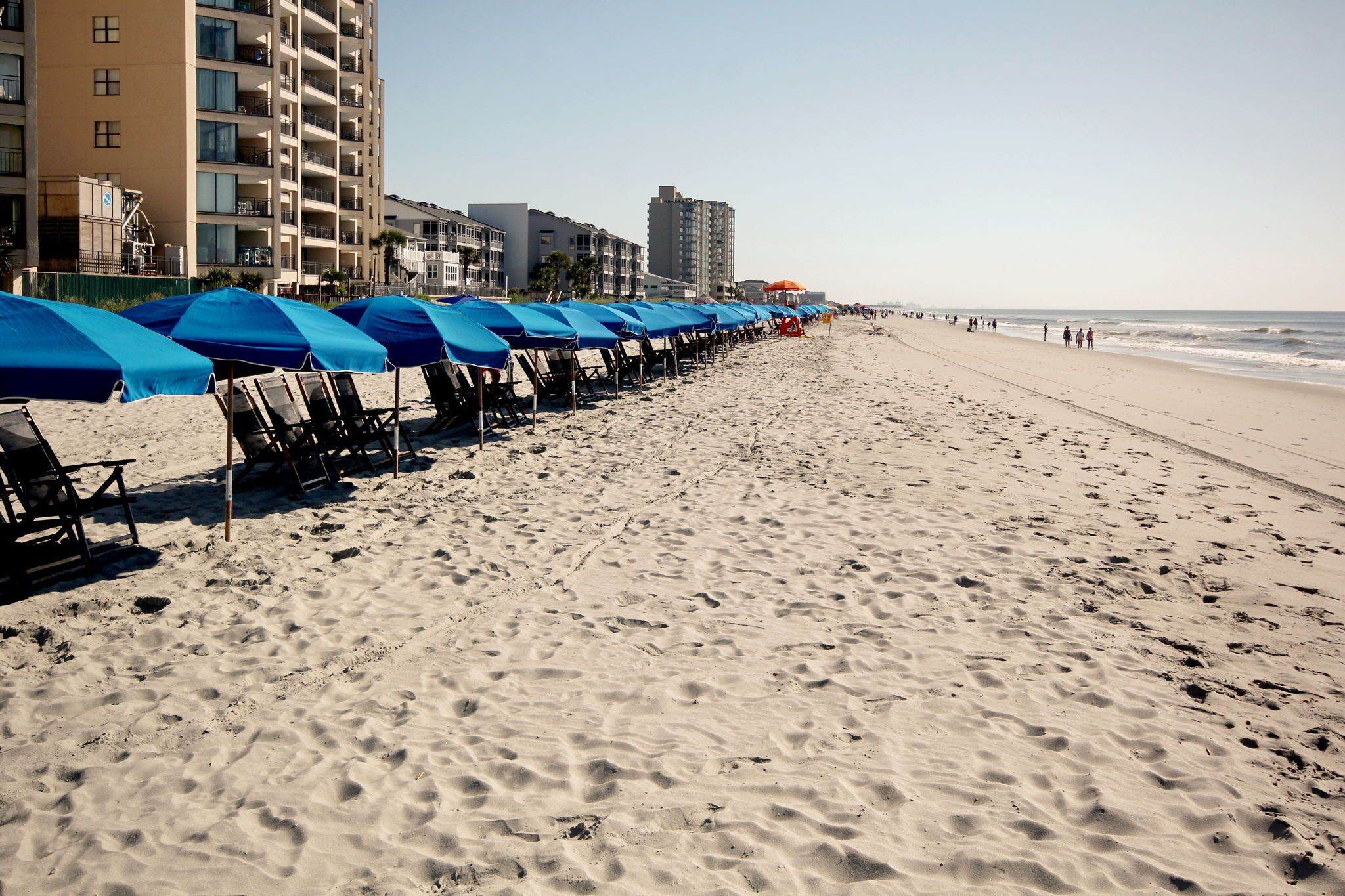 A beach with rows of blue umbrellas and beach chairs, a sandy shore, and buildings in the background.