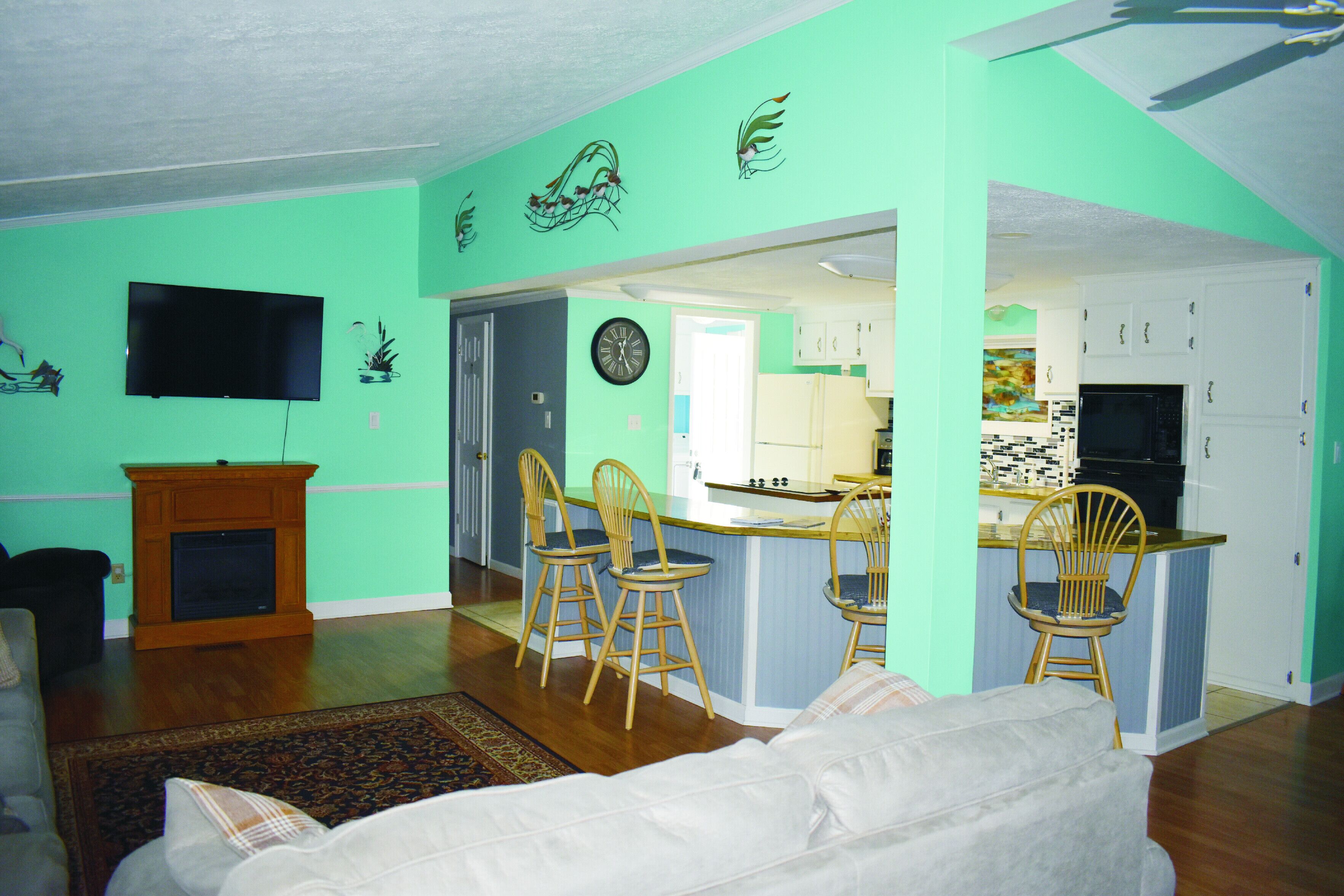A living room with a sofa, a TV, and a fireplace. There is a kitchen with a microwave, cabinets, and a dining area with chairs.