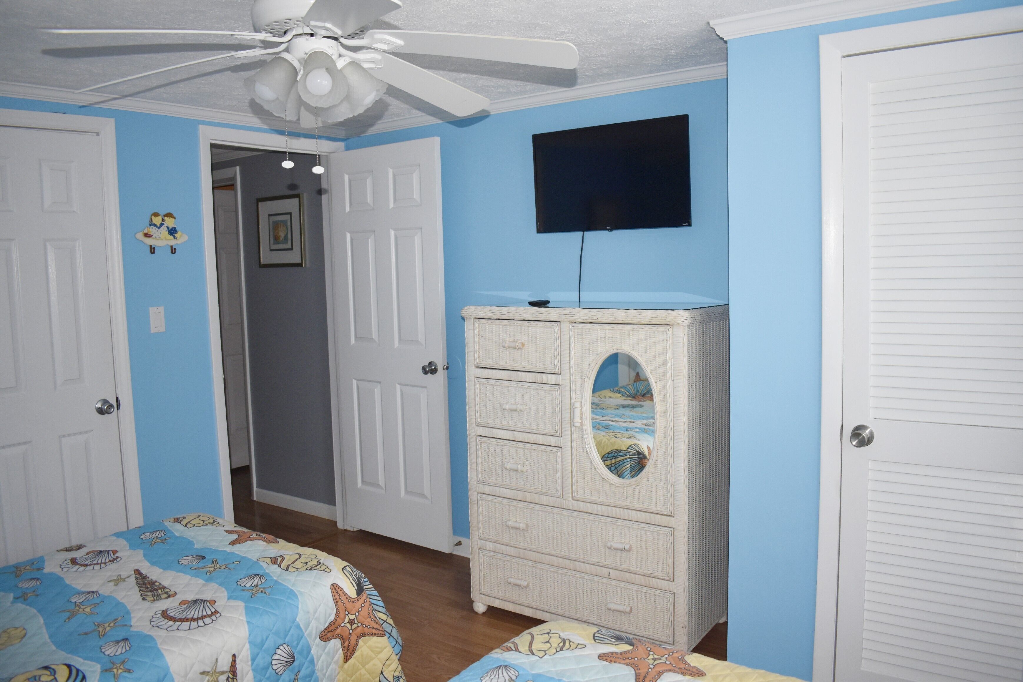 A bedroom with a bed, a dresser, a ceiling fan, and a TV on the wall.