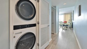 Un cuarto de lavandería con dos lavadoras, una puerta cerrada, y un comedor con mesa y sillas al fondo.
