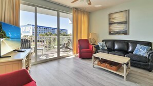Una sala de estar moderna con un sofá de cuero, un sillón rojo, una mesa de centro de madera y un televisor sobre un mueble. Se observa un ventilador de techo, y hay un ventanal con vistas a un edificio.