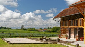 A rustic building with a red-tiled roof and wooden balcony, surrounded by a well-maintained garden and fields.