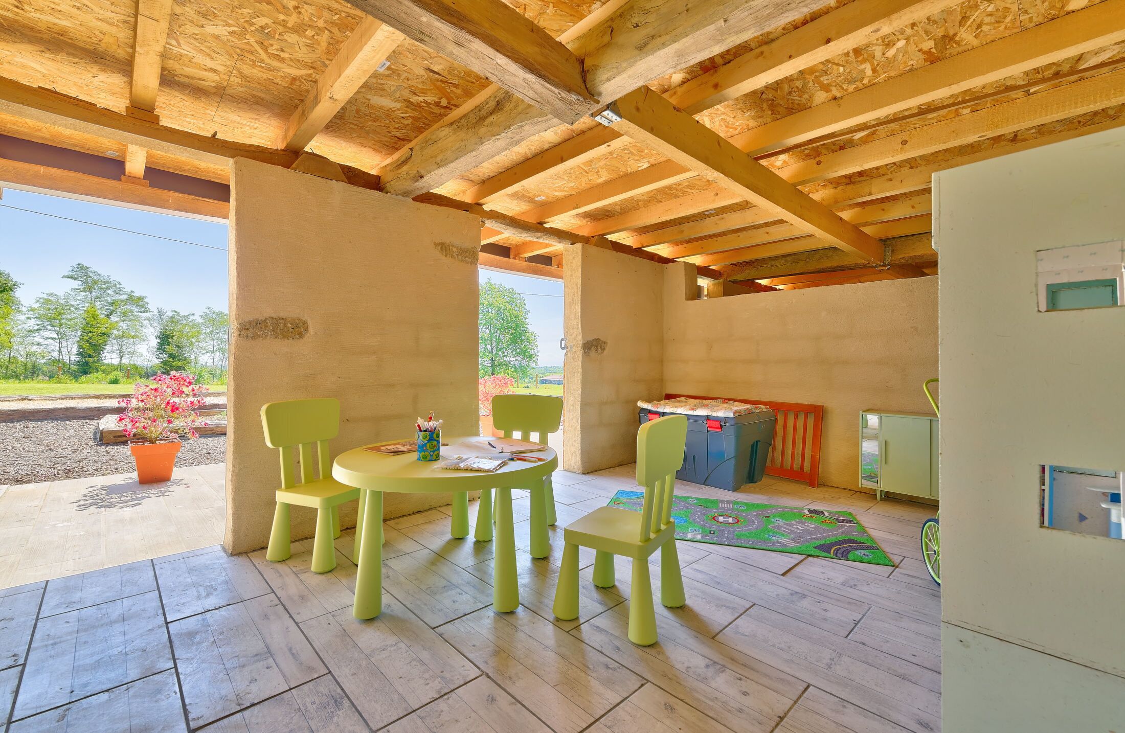 A covered patio area with a wooden ceiling, a table with chairs, a small play area, and a view of the outdoors.
