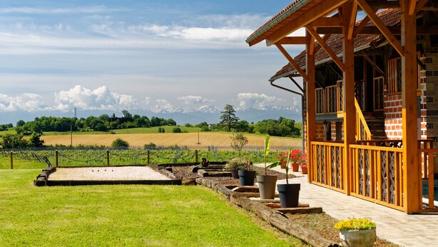 A wooden building with a balcony overlooking a landscaped garden and a distant mountain range. - Charming 9-person gîte with heated swimming pool (Peyrusse-Grande)