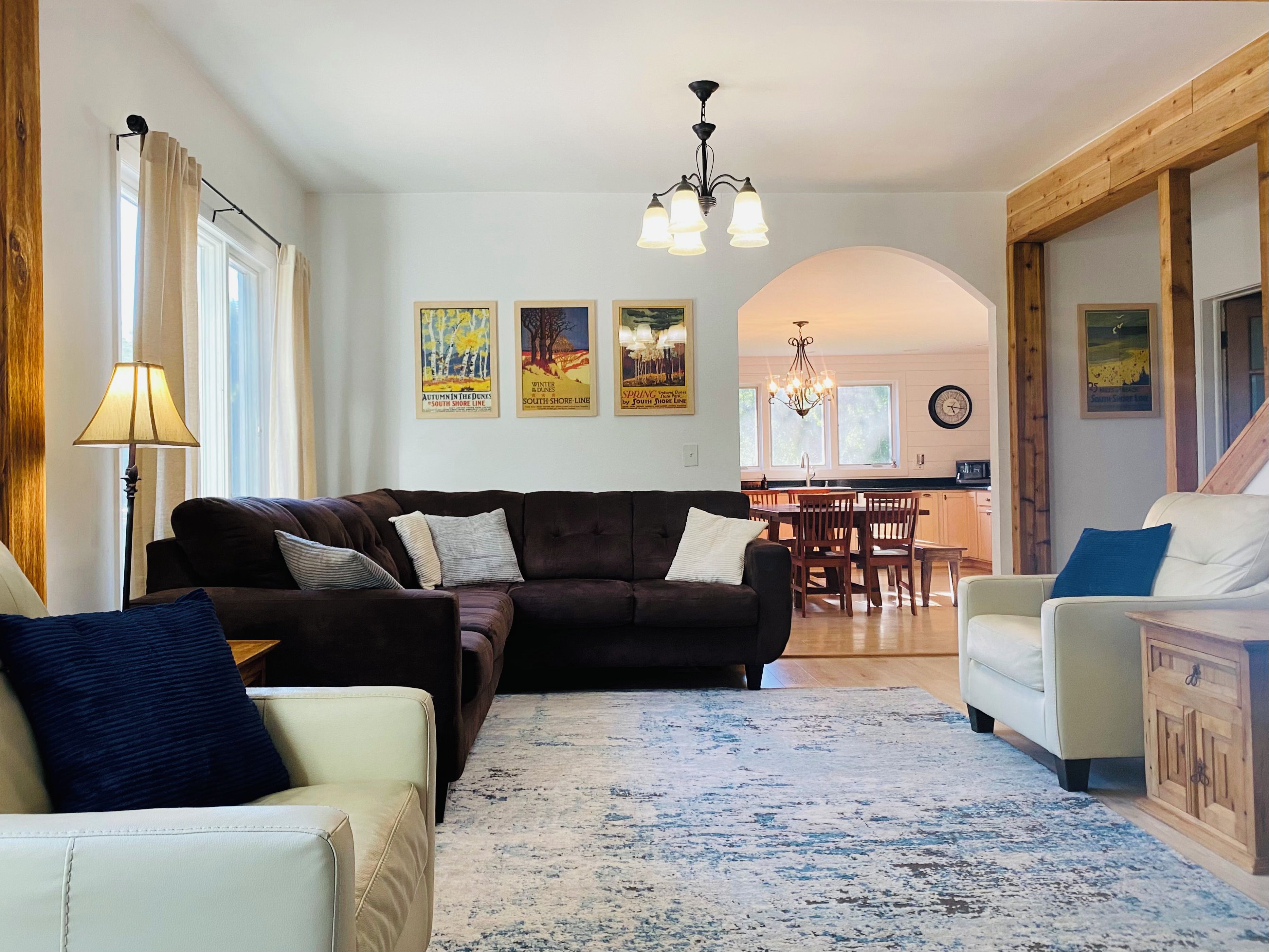 Uma sala de estar com sofá, poltronas, tapete e um espaço de jantar com mesa e cadeiras. Há quadros pendurados na parede e um lustre.
