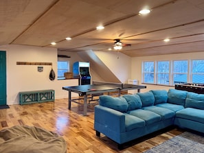 Una habitación con mesa de ping pong, sofá azul, mesa de fútbol de mesa, ventilador de techo y ventana con vistas.
