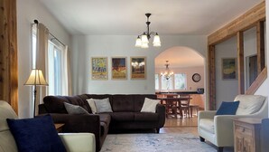 A living room with a brown sofa, a white sofa, and a beige armchair. There is a dining area with a wooden table and chairs. The room has wooden beams on the ceiling and a chandelier.