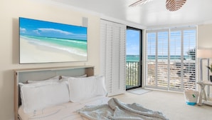 Une chambre avec un lit, une grande fenêtre donnant sur la plage et un ventilateur de plafond.