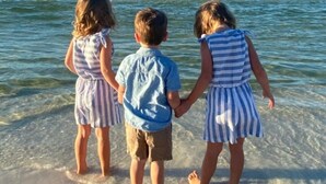 Trois enfants se tenant la main sur une plage, avec une eau claire et une mer calme en arrière-plan.