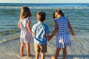 Drei Kinder halten Händchen am Strand, im Hintergrund ruhiges Wasser und ein stilles Meer.