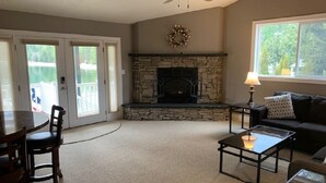 A living room with a stone fireplace, a ceiling fan, a dining table with chairs, a sofa, and a window with a view of greenery. - Waterfront Home-Great Boating/Fishing/Beach. Mountain Views, close to Sandpoint (Sagle)