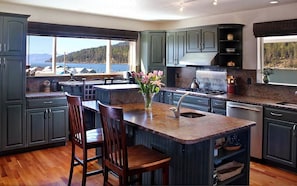 A modern kitchen with dark green cabinets, a central island with bar stools, a view of the lake, and a stove.