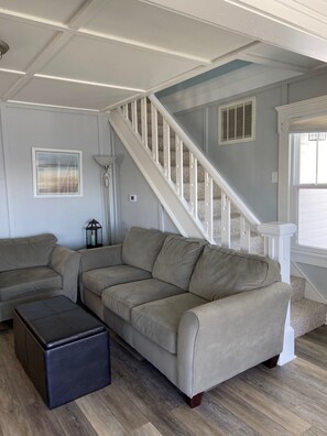 A living room with a beige sectional sofa, a dark ottoman, a floor lamp, and a staircase with white railings.