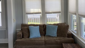 Ein Zimmer mit einem braunen Sofa, zwei blauen Kissen, einem Weidenhocker und einem Deckenventilator.