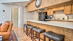 Une cuisine moderne avec des meubles en bois, une cuisinière encastrée et une grande horloge. Il y a trois tabourets au comptoir et un canapé dans le salon attenant.