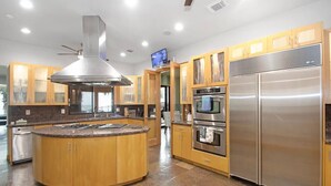 A modern kitchen with a central island, stainless steel appliances, wooden cabinets, and a ceiling fan. - Luxury Waterfront Estate w/ Heated Pools, Private Pier & Walk to Boardwalk (Kemah)