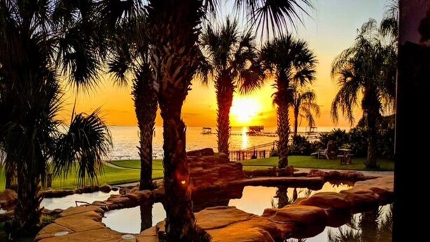 A sunset view with palm trees, a pool, and a pier. - Luxury Waterfront Estate w/ Heated Pools, Private Pier & Walk to Boardwalk (Kemah)