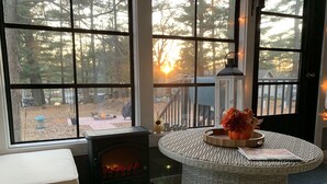 Ein gemütliches Zimmer mit Kamin, einem Korbstuhl mit Vase und Blick auf einen Wald durch große Fenster.