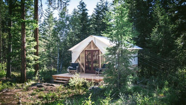 A rustic cabin with a wooden deck and a large front door, surrounded by dense forest. - A Family Glamping Adventure Awaits (Victor)