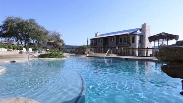 A swimming pool with a wooden deck, lounge chairs, and a building with a large window and a chimney.