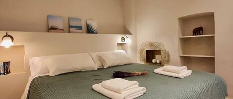 A neatly made bed with a green blanket, two pillows, and folded towels on a textured bedspread. There are wall-mounted shelves with decorative items and a wall-mounted lamp.