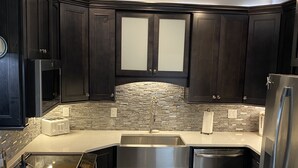 A modern kitchen with dark cabinetry, a stone backsplash, stainless steel appliances, and a light-colored countertop.