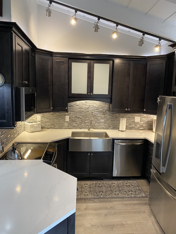 A modern kitchen with dark cabinetry, a stone backsplash, stainless steel appliances, and a light-colored countertop.